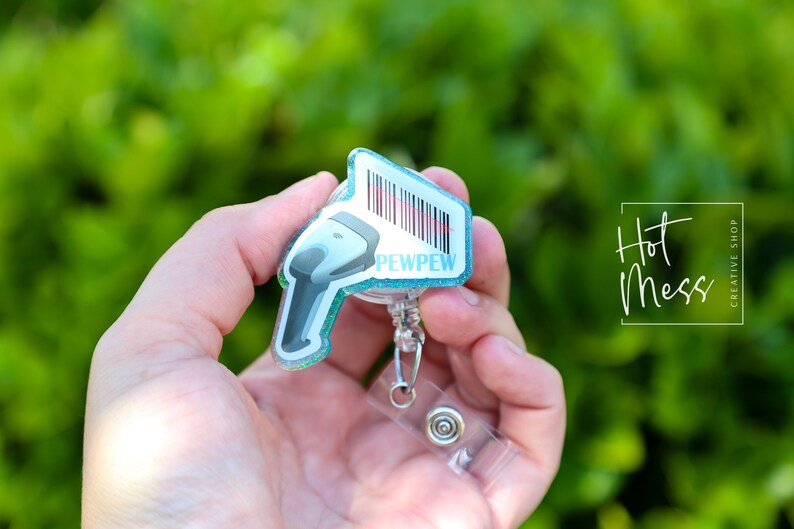 May include: A teal blue glitter badge holder with a white scanner design and the text "PEWPEW". The badge holder is attached to a clear plastic retractable badge reel.