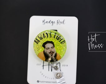 May include: A green glitter badge reel featuring a picture of a man with the text "Always Tired" on it. The badge reel is displayed on a white card with the text "Hot Mess Creative Shop" on it.