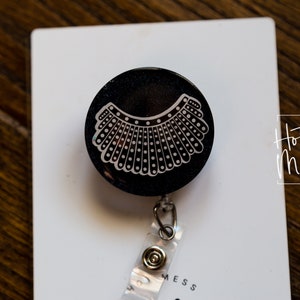 May include: Black retractable badge holder with a white graphic of a judge's collar. The badge holder is on a white background with the text "Hot Mess Creative Shop".