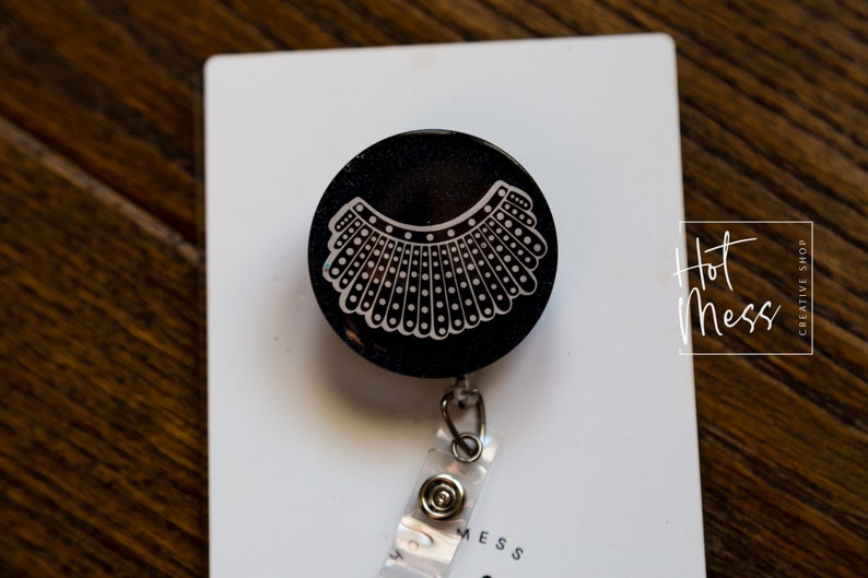 May include: Black retractable badge holder with a white graphic of a judge's collar. The badge holder is on a white background with the text "Hot Mess Creative Shop".