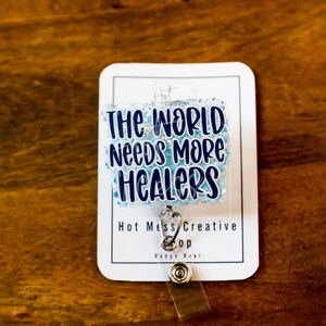 May include: A white badge reel with the text "THE WORLD Needs MORE HEALERS" in blue lettering. The badge reel has a silver clip and a clear plastic strap. The background is a dark brown wooden surface.