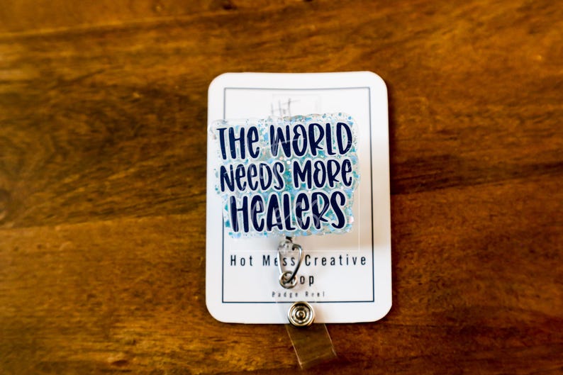 May include: A white badge reel with the text "THE WORLD Needs MORE HEALERS" in blue lettering. The badge reel has a silver clip and a clear plastic strap. The background is a dark brown wooden surface.