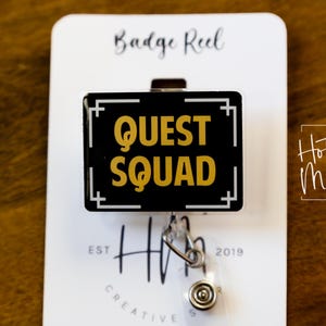 May include: A black and gold badge reel with the words "QUEST SQUAD" in gold lettering. The badge reel is rectangular with a white border and a silver clip. The background is a wooden surface.
