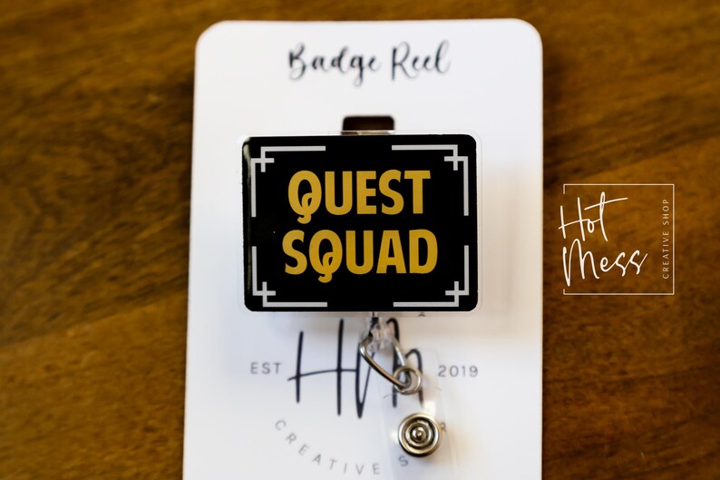 May include: A black and gold badge reel with the words "QUEST SQUAD" in gold lettering. The badge reel is rectangular with a white border and a silver clip. The background is a wooden surface.