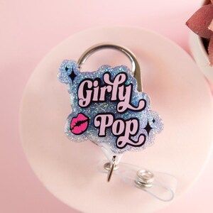 Op de afbeelding: Een badge reel met de woorden "Girly Pop" in roze en zwarte letters, geaccentueerd met een roze lip-afbeelding. De badge reel heeft een glitterende blauwe achtergrond en een zilveren clip.