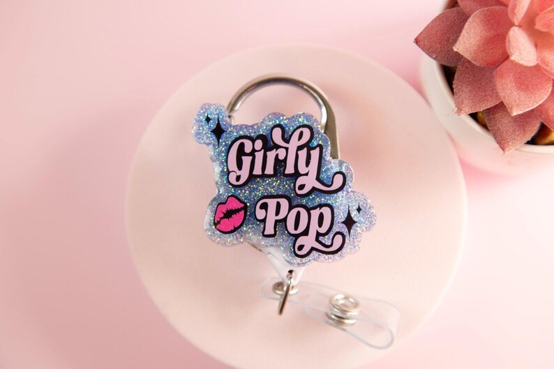 Op de afbeelding: Een badge reel met de woorden "Girly Pop" in roze en zwarte letters, geaccentueerd met een roze lip-afbeelding. De badge reel heeft een glitterende blauwe achtergrond en een zilveren clip.