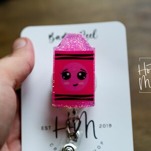 May include: Pink glitter and black striped crayon-shaped retractable badge reel with a cute cartoon face. The reel is attached to a white card with the text "Hot Mess Creative Shop" and "Est. 2019".