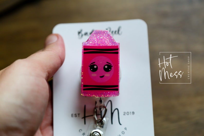 May include: Pink glitter and black striped crayon-shaped retractable badge reel with a cute cartoon face. The reel is attached to a white card with the text "Hot Mess Creative Shop" and "Est. 2019".