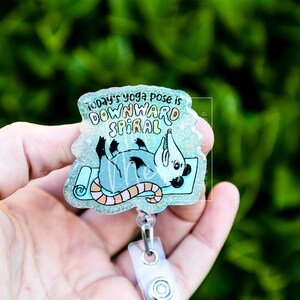 Op de afbeelding: Een badge reel met een cartoon opossum in een yogahouding op een blauwe mat. De tekst luidt "Today's yoga pose is downward spiral". De badge reel heeft een doorzichtige plastic behuizing en een witte clip.