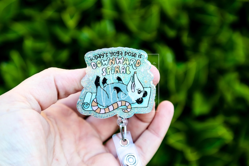 Op de afbeelding: Een badge reel met een cartoon opossum in een yogahouding op een blauwe mat. De tekst luidt "Today's yoga pose is downward spiral". De badge reel heeft een doorzichtige plastic behuizing en een witte clip.