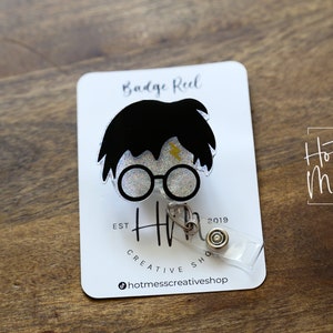 Puede incluir: Un carrete de credencial blanco con un dise&ntilde;o de personaje de Harry Potter en blanco y negro. El personaje tiene una cicatriz de rayo en la frente y lleva gafas redondas. El texto "Badge Reel" est&aacute; impreso en la tarjeta detr&aacute;s del personaje. El texto "Hot Mess Creative Shop" est&aacute; impreso en la tarjeta.