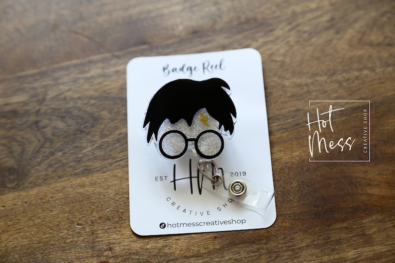 Puede incluir: Un carrete de credencial blanco con un dise&ntilde;o de personaje de Harry Potter en blanco y negro. El personaje tiene una cicatriz de rayo en la frente y lleva gafas redondas. El texto "Badge Reel" est&aacute; impreso en la tarjeta detr&aacute;s del personaje. El texto "Hot Mess Creative Shop" est&aacute; impreso en la tarjeta.
