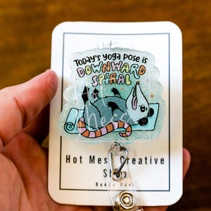 Op de afbeelding: Een badge reel met een helder, glinsterend acrylontwerp. Het ontwerp toont een cartoon opossum op een yogamat met de tekst "Today's yoga pose is DOWNWARD SPIRAL". De badge reel heeft een metalen clip en een intrekbaar koord.
