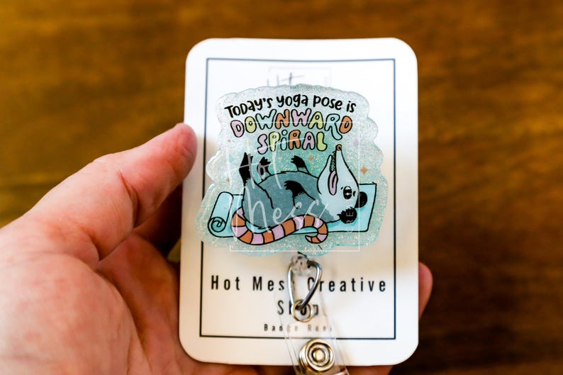 Op de afbeelding: Een badge reel met een helder, glinsterend acrylontwerp. Het ontwerp toont een cartoon opossum op een yogamat met de tekst "Today's yoga pose is DOWNWARD SPIRAL". De badge reel heeft een metalen clip en een intrekbaar koord.