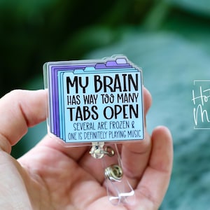 May include: A clear badge reel with a design of stacked file tabs. The text reads "MY BRAIN HAS WAY TOO MANY TABS OPEN. SEVERAL ARE FROZEN & ONE IS DEFINITELY PLAYING MUSIC." The badge reel has a silver clip and a clear plastic strap.