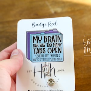 May include: A white badge reel with the text "MY BRAIN HAS WAY TOO MANY TABS OPEN" in black and blue. The badge reel has a design of file folders in purple and blue.