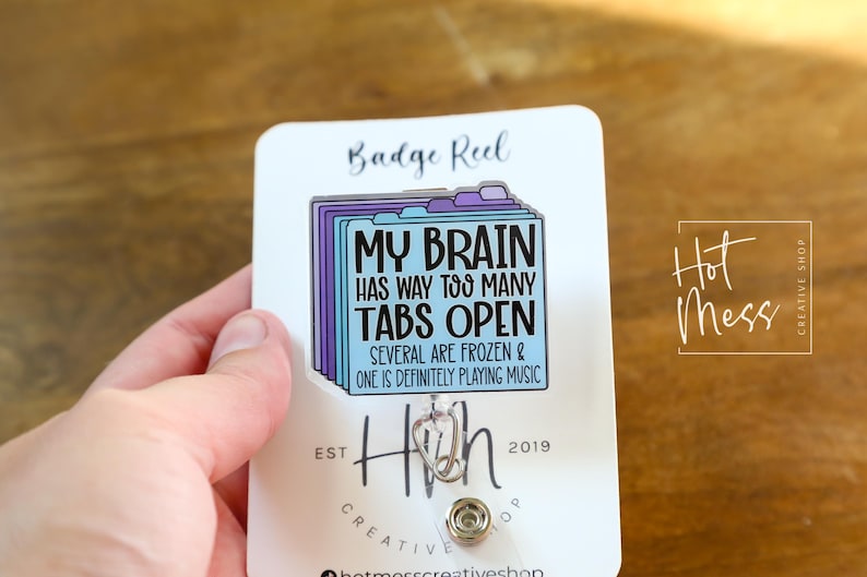 May include: A white badge reel with the text "MY BRAIN HAS WAY TOO MANY TABS OPEN" in black and blue. The badge reel has a design of file folders in purple and blue.