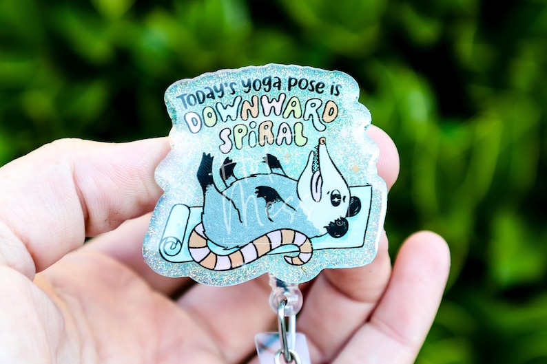 Op de afbeelding: Een badge met een cartoon opossum op een yogamat. De tekst luidt "Today's yoga pose is downward spiral" in een speels lettertype. De badge heeft een glitterende, iriserende afwerking en een intrekbare rol.