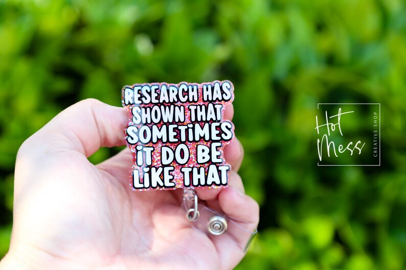 Op de afbeelding: Een roze glitter badgehouder met een witte tekstoverlay die "Research has shown that sometimes it do be like that" luidt.