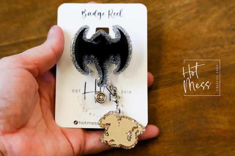 Puede incluir: Un carrete de insignia plateado con un dise&ntilde;o de drag&oacute;n negro con purpurina. El carrete est&aacute; unido a un amuleto de purpurina dorada con un dise&ntilde;o de drag&oacute;n. El carrete est&aacute; etiquetado como "Badge Reel" y "Hot Mess Creative Shop".