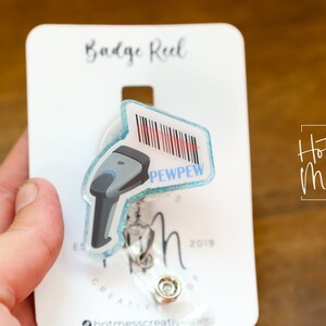 May include: A blue and white badge reel with a clear plastic cover. The reel has a barcode scanner design with the text "PEWPEW" on it. The reel is attached to a white card with the text "Badge Reel" and "Hot Mess Creative Shop".