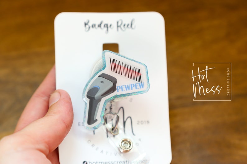 May include: A blue and white badge reel with a clear plastic cover. The reel has a barcode scanner design with the text "PEWPEW" on it. The reel is attached to a white card with the text "Badge Reel" and "Hot Mess Creative Shop".