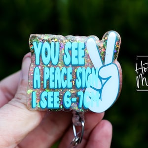 Puede incluir: Un portatarjetas brillante con el texto "YOU SEE A PEACE SIGN I SEE 6-7". El diseño incluye un signo de la paz y está en tonos azules y blancos. El portatarjetas tiene un clip plateado y las palabras "Hot Mess Creative Shop".