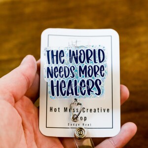 May include: A white badge reel with the text "THE WORLD NEEDS MORE HEALERS" in blue lettering. The badge has a clear, glittery overlay and a silver-colored clip. The words "Hot Mess Creative Shop" are printed at the bottom.
