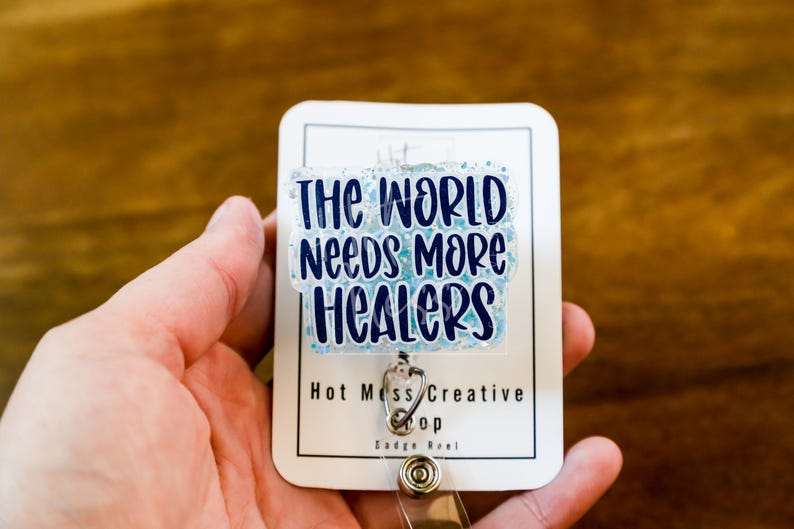 May include: A white badge reel with the text "THE WORLD NEEDS MORE HEALERS" in blue lettering. The badge has a clear, glittery overlay and a silver-colored clip. The words "Hot Mess Creative Shop" are printed at the bottom.