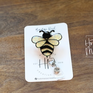 May include: A badge reel featuring a black and yellow bee design with glitter accents. The reel is attached to a white card with the text "Badge Reel" and "Hot Mess Creative Shop". The background is a wooden surface.