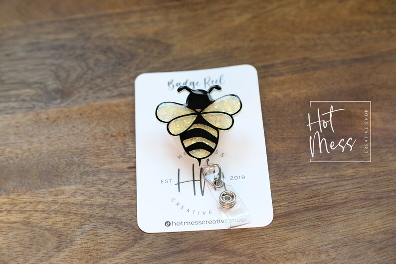 May include: A badge reel featuring a black and yellow bee design with glitter accents. The reel is attached to a white card with the text "Badge Reel" and "Hot Mess Creative Shop". The background is a wooden surface.