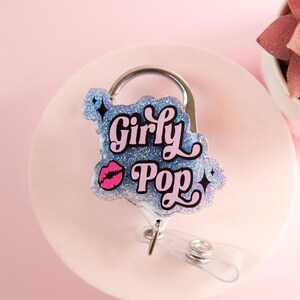 Op de afbeelding: Een glitter badge reel met de woorden "Girly Pop" in een speels lettertype, met een roze lip afbeelding. De badge reel heeft een zilveren clip en een transparant intrekbaar koord, tegen een zachtroze achtergrond.