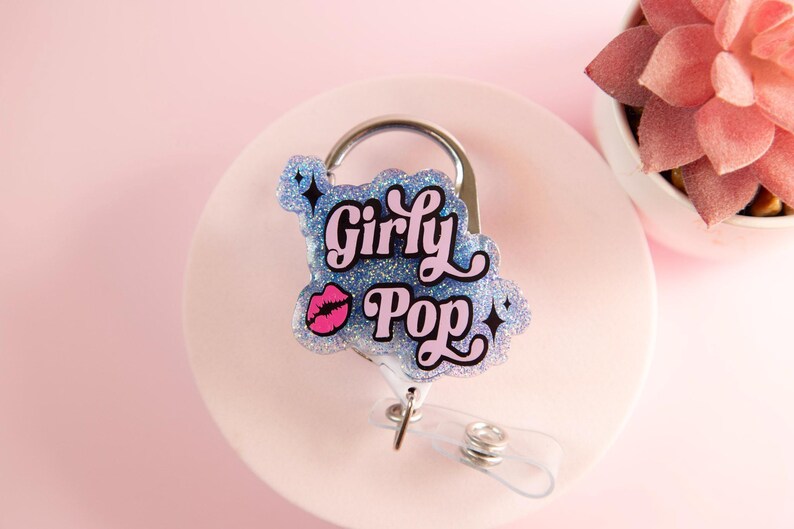 Op de afbeelding: Een glitter badge reel met de woorden "Girly Pop" in een speels lettertype, met een roze lip afbeelding. De badge reel heeft een zilveren clip en een transparant intrekbaar koord, tegen een zachtroze achtergrond.