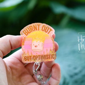 May include: A glittery badge reel with the text "BURNT OUT BUT OPTIMISTIC" in a pink and yellow design. The badge reel features a cartoon marshmallow character and the words "Hot Mess Creative Shop".