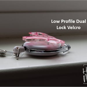 May include: A pink and white retractable badge holder with a low profile dual lock velcro closure. The holder features a pink glitter design with a heart and the text "Hot Mess" in black script. The holder is attached to a clear plastic strap with a metal clip.