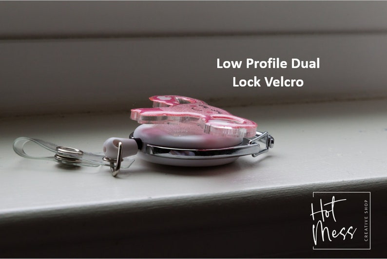 May include: A pink and white retractable badge holder with a low profile dual lock velcro closure. The holder features a pink glitter design with a heart and the text "Hot Mess" in black script. The holder is attached to a clear plastic strap with a metal clip.