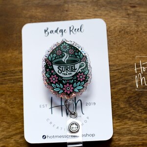 May include: A teal, pink, and white badge reel with a floral design and a teacup with the text "SURIEL TEA CO."  The badge reel is attached to a clear plastic retractable cord.