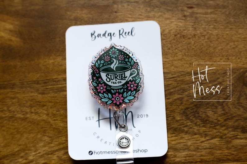 May include: A teal, pink, and white badge reel with a floral design and a teacup with the text "SURIEL TEA CO."  The badge reel is attached to a clear plastic retractable cord.