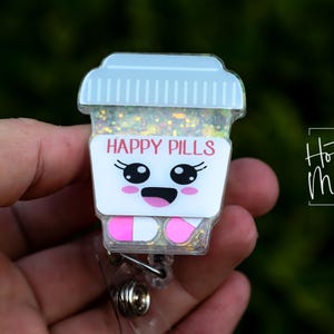 May include: A novelty badge reel with a cartoon coffee cup design. The cup has the words "HAPPY PILLS" in pink, a smiling face, and pink and white pill-shaped feet. The cup is filled with glitter.