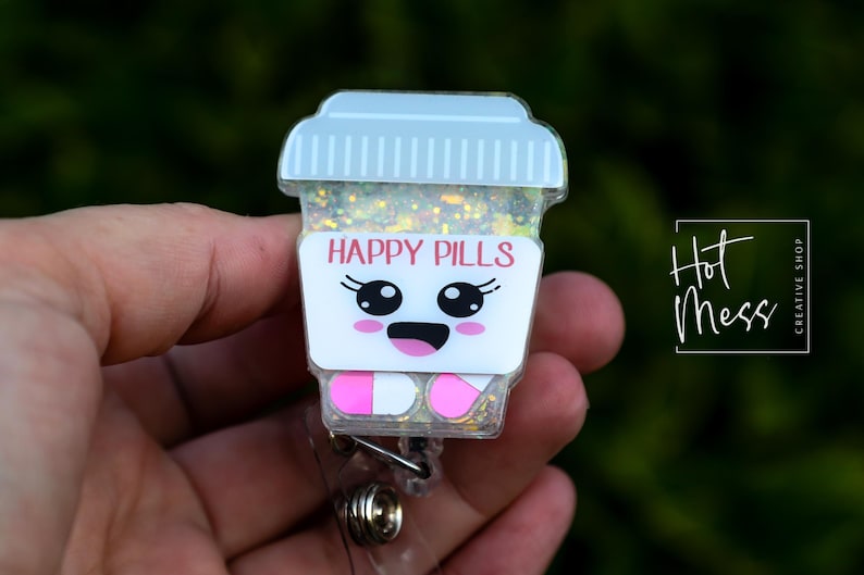 May include: A novelty badge reel with a cartoon coffee cup design. The cup has the words "HAPPY PILLS" in pink, a smiling face, and pink and white pill-shaped feet. The cup is filled with glitter.
