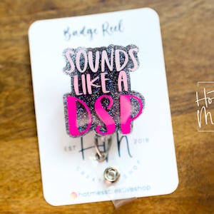 Könnte beinhalten: Ein silberner Glitzer-Badge-Rollenhalter mit einem pinkfarbenen und weißen Design "Sounds Like a DSP". Der Rollenhalter ist an einer weißen Karte befestigt, auf der der Text "Hot Mess Creative Shop" gedruckt ist.