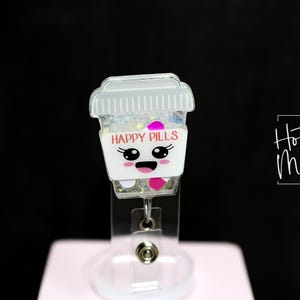 May include: A badge reel featuring a cartoon coffee cup design. The cup is white with the words "HAPPY PILLS" and a smiling face. The cup is filled with glitter and pink accents. The reel has a clear plastic base.
