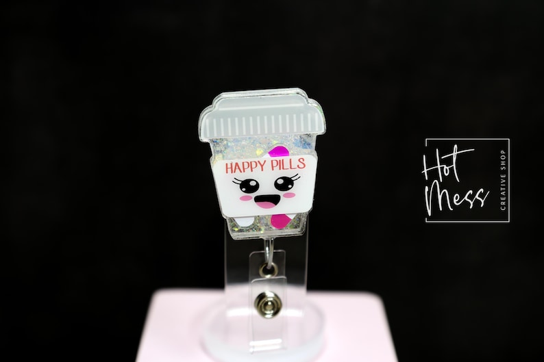 May include: A badge reel featuring a cartoon coffee cup design. The cup is white with the words "HAPPY PILLS" and a smiling face. The cup is filled with glitter and pink accents. The reel has a clear plastic base.