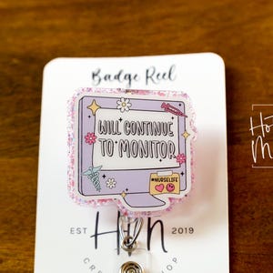 May include: A badge reel with a computer-shaped design, featuring the text "WILL CONTINUE TO MONITOR." The design includes flowers, stars, and medical symbols. The reel is on a white card with the text "Badge Reel" and "Hot Mess Creative Shop."