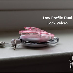 May include: A pink and white retractable badge holder with a low profile dual lock velcro closure. The holder features a pink glitter design with a heart and the text "Hot Mess" in black script. The holder is attached to a clear plastic strap with a metal clip.