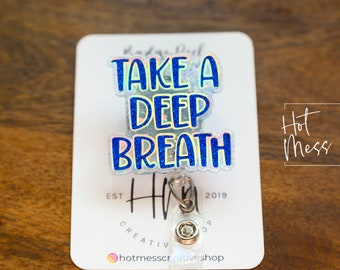 以下が含まれることがあります： 「Take a Deep Breath」と書かれた青と白の虹色のグリッターステッカーが付いたシルバーのリトラクタブルバッジホルダー。ステッカーは、「Hot Mess Creative Shop」と「Est 2019」の文字が書かれた白いカードに貼られています。