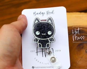 May include: A black glitter bat badge reel with a white outline and a silver retractable badge holder. The badge reel is attached to a white card with the text "Badge Reel", "Hot Mess Creative Shop", "Est 2019", and "@hotmesscreativeshop".