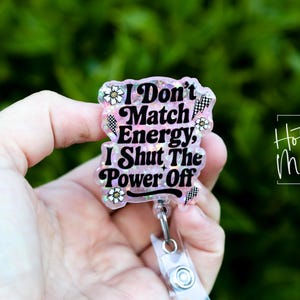 May include: A clear, rectangular badge reel with the text "I Don't Match Energy, I Shut The Power Off." The badge has a pink and white background with floral and checkered accents. The words are in black.
