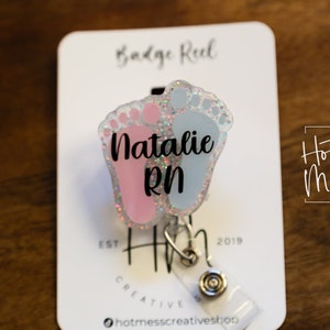 May include: A pink and blue glitter baby feet badge reel with the name "Natalie RN" printed on it. The badge reel is attached to a white card with the text "Badge Reel" and "Hot Mess Creative Shop" printed on it.