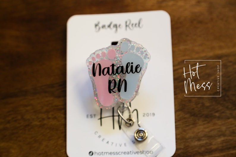 May include: A pink and blue glitter baby feet badge reel with the name "Natalie RN" printed on it. The badge reel is attached to a white card with the text "Badge Reel" and "Hot Mess Creative Shop" printed on it.